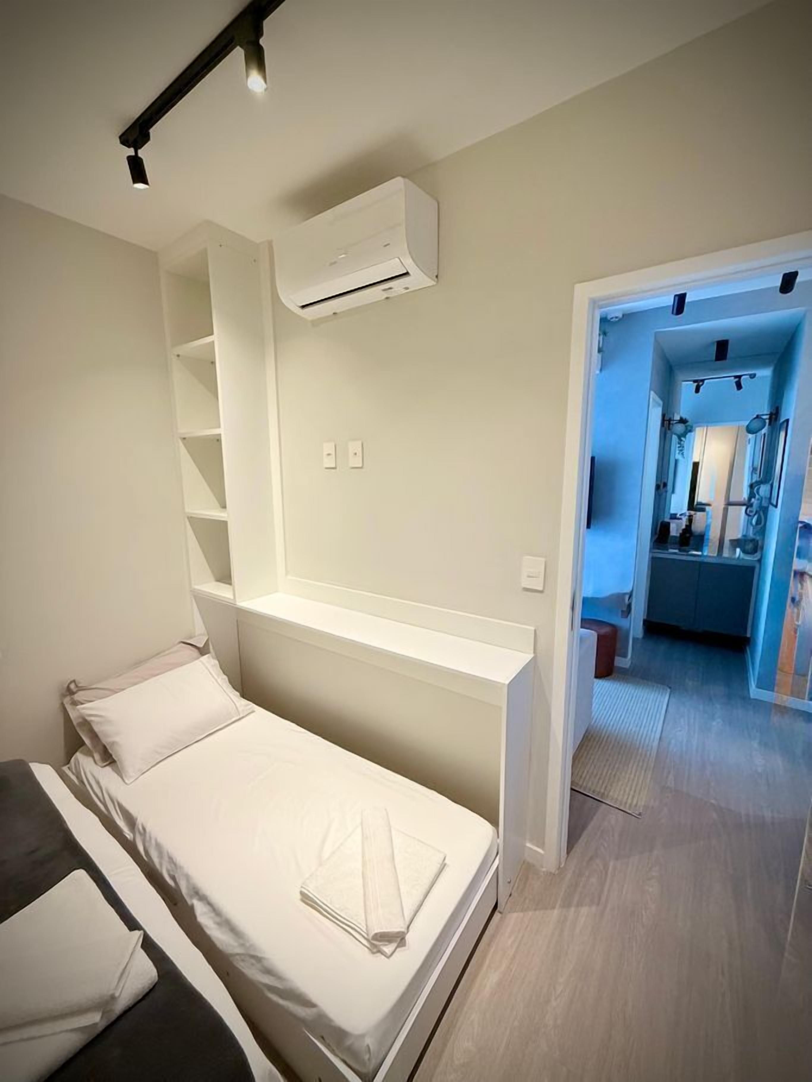 A bedroom with a bed, a built-in shelf, an air conditioning unit, and a bathroom visible through an open door.