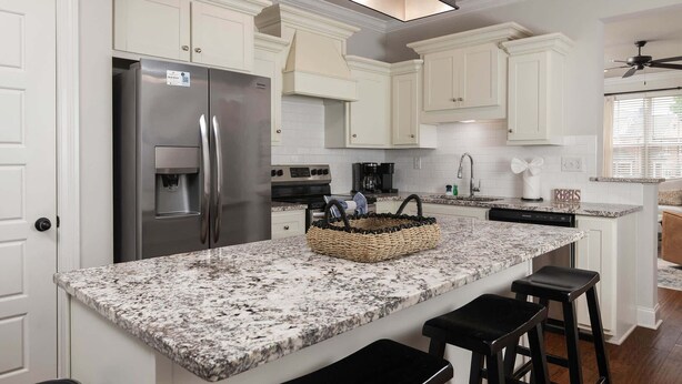 Una cocina con gabinetes blancos, encimera de granito, refrigerador de acero inoxidable, cafetera y lavavajillas.