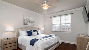 Un dormitorio con cama, mesitas de noche, ventilador de techo, ventana con persianas y televisor instalado en la pared.