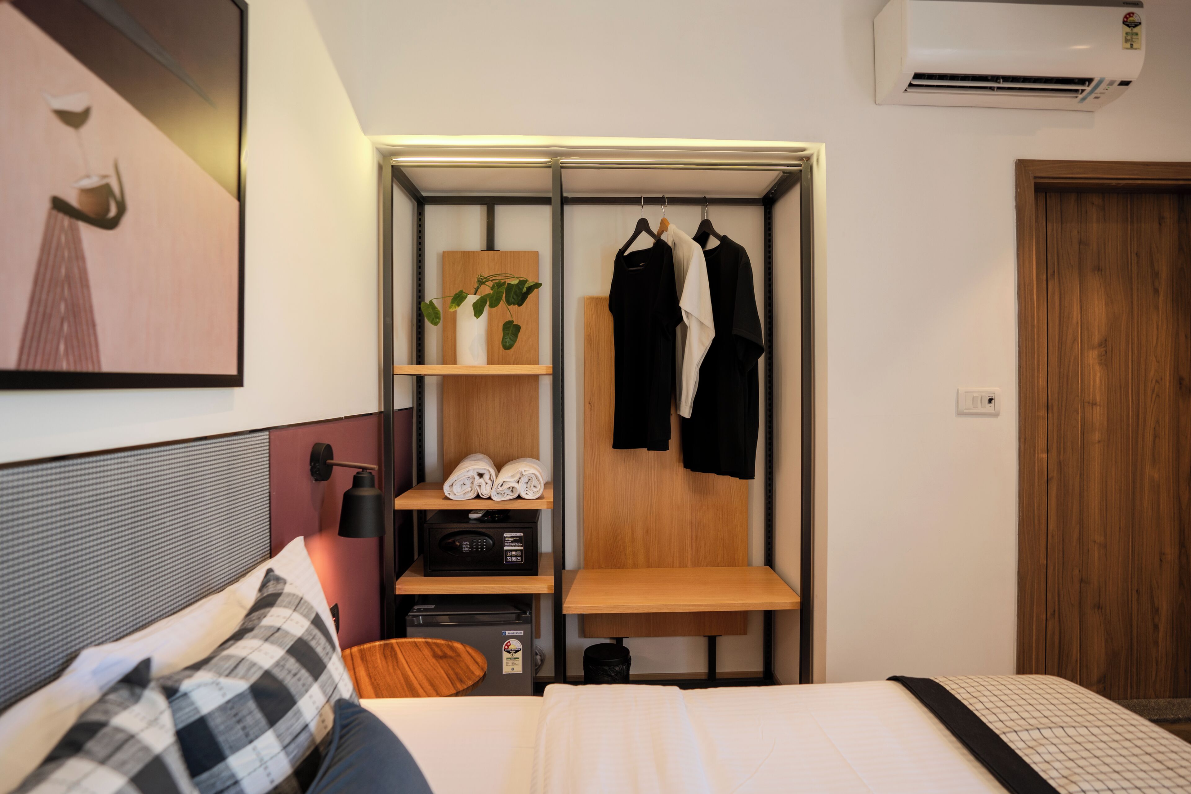 A hotel room with a bed, a desk with a computer, a shelf with towels, a hanging coat rack, and an air conditioning unit.