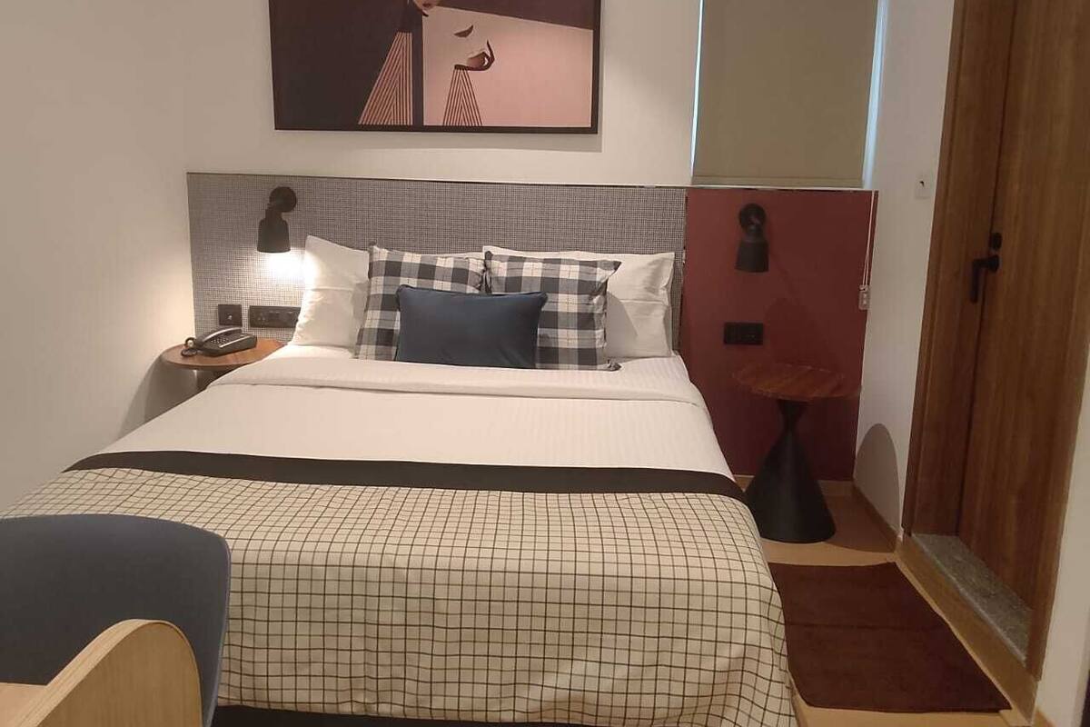 A hotel room with a bed, a nightstand with a lamp, a chair, a small table, and a door.
