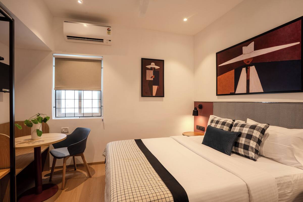 Una habitación de hotel moderna con una cama, un escritorio con una silla, una ventana con persianas y un cuadro en la pared.