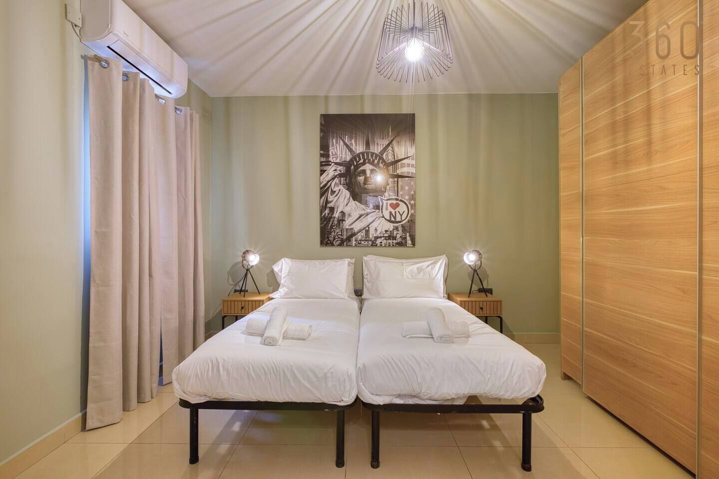 Una cama doble con ropa de cama blanca, dos lámparas de noche, un cuadro de un paisaje urbano en la pared y un armario de madera.