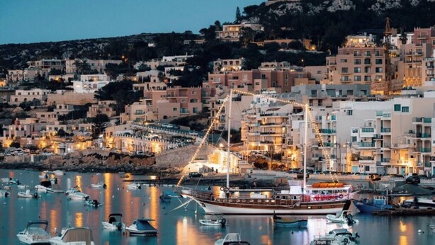 A coastal town at dusk with a marina filled with boats, buildings with balconies, and a prominent hill in the background.