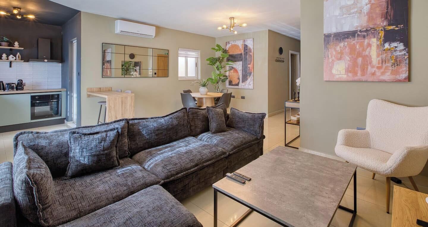Una sala moderna con un sofá modular, una mesa de centro y un sillón blanco. Cuenta con una zona de cocina, una mesa de comedor y sillas, un espejo grande y una planta en maceta.