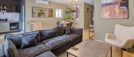 Una sala moderna con un sofá modular, una mesa de centro y un sillón blanco. Cuenta con una zona de cocina, una mesa de comedor y sillas, un espejo grande y una planta en maceta.