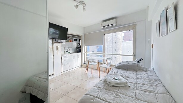 Quarto compacto com cama, pequena cozinha, televisão e janela com vista.