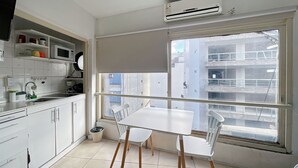 Uma cozinha pequena e limpa, com armários brancos, uma mesa branca e cadeiras. Há uma janela com vista para edifícios.