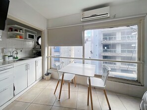 Eine kleine, saubere Küche mit weißen Schränken, einem weißen Tisch und Stühlen. Ein Fenster bietet Blick auf umliegende Gebäude.