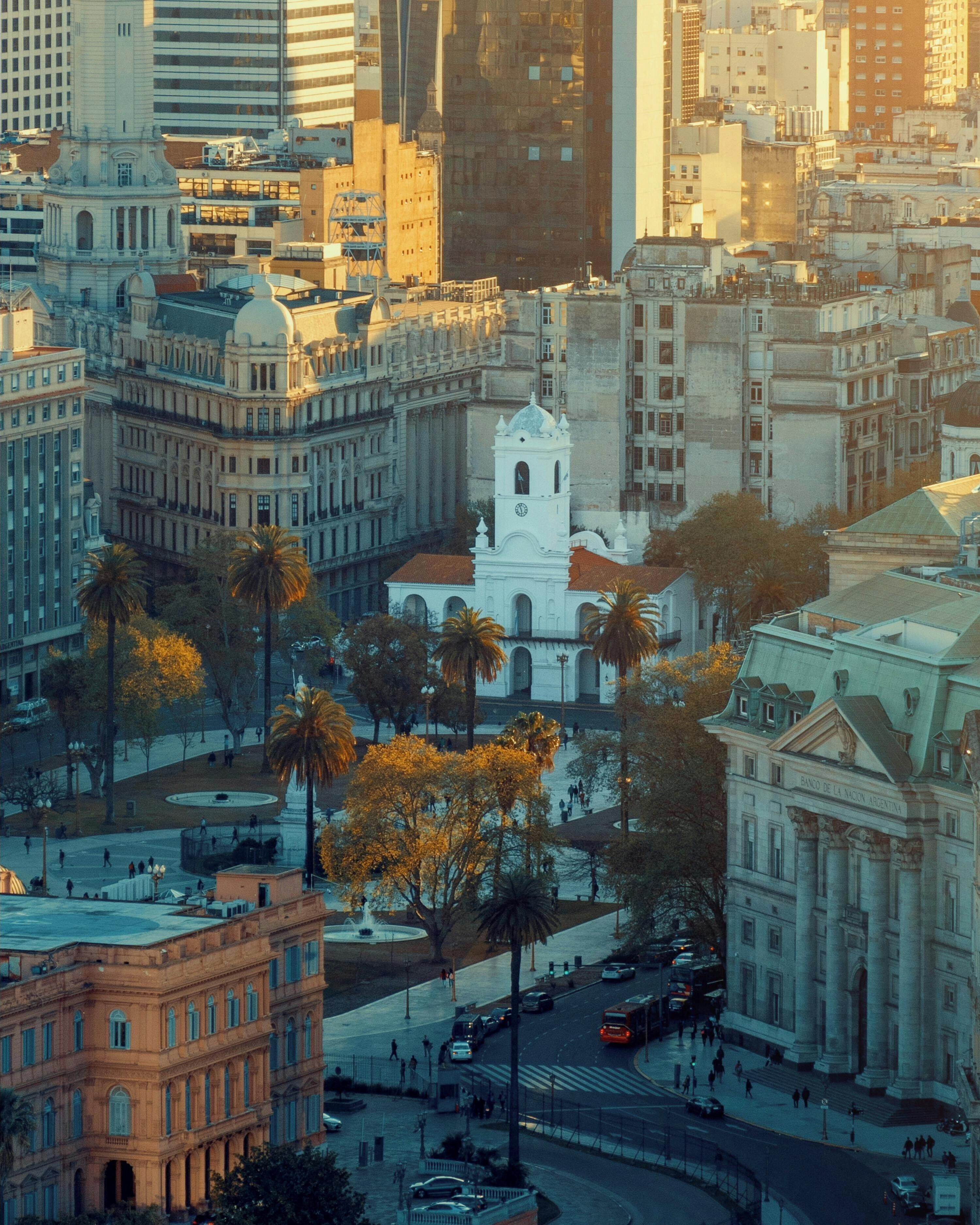 Paisagem urbana com arquitetura histórica, incluindo um edifício branco de destaque com torre do relógio, cercado por palmeiras e edifícios modernos.