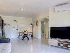 Un saló amb televisió, una zona de menjador amb taula i cadires, una porta blanca i un passadís que condueix a un bany.