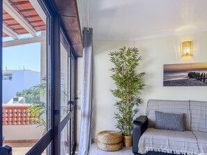 Habitació amb vistes a través d'una porta de vidre, una planta de bambú, una cistella teixida, un sofà amb una manta i una làmpada penjada a la paret.