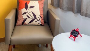 Une pièce avec un fauteuil gris, une table ronde blanche, un miroir hexagonal et un petit tableau noir sur lequel est écrit « Bem Vindo ».