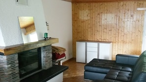 A living room with a fireplace, a sofa, a small kitchen area, and wooden walls. - Haus Gonzenblick, (Flumserberg Tannenheim), by Interhome (Flumserberg Tannenheim)