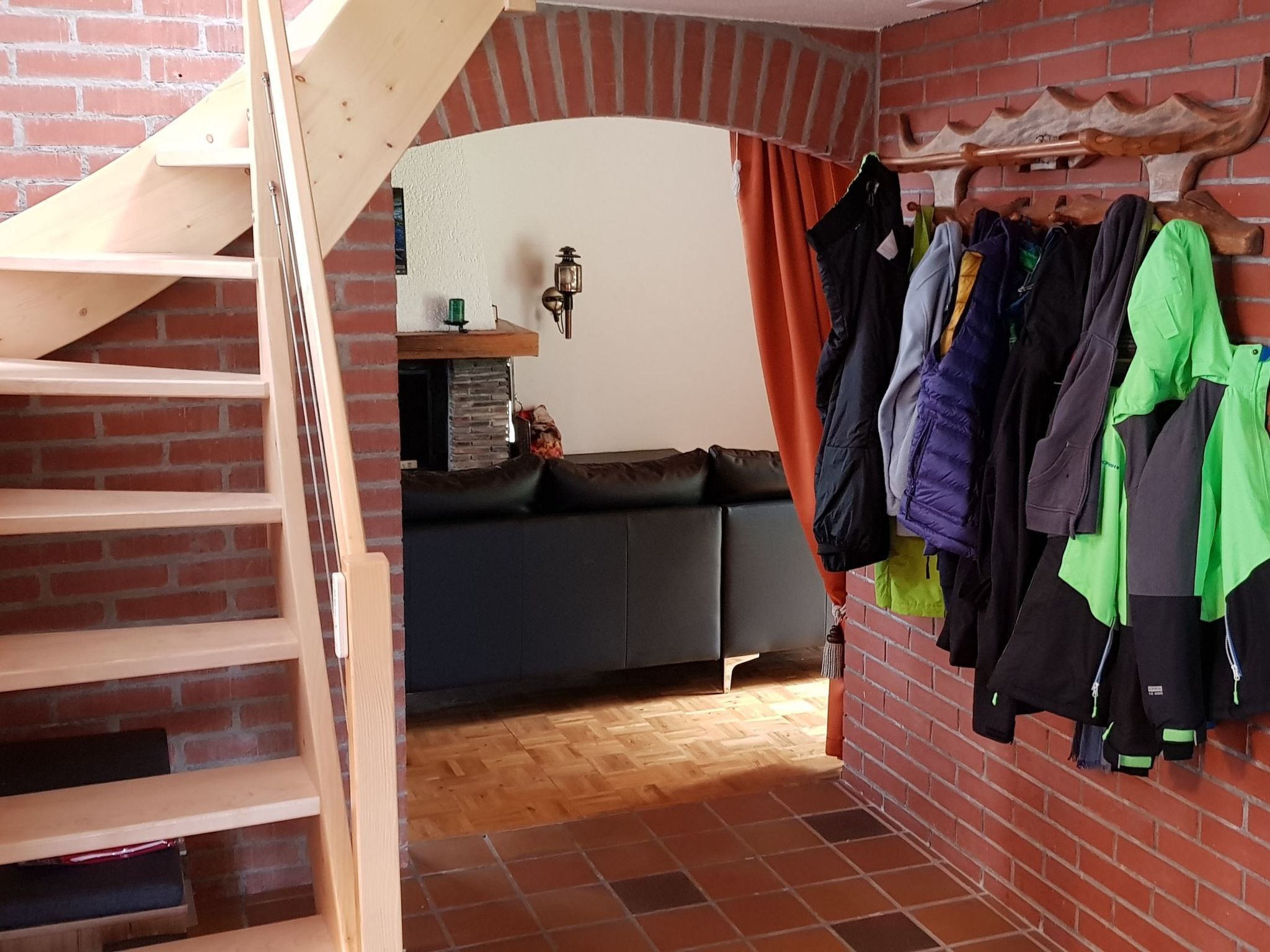 Une pièce avec un mur en briques, un escalier en bois, un canapé gris et un portant à vêtements sur lequel sont accrochées différentes vestes.