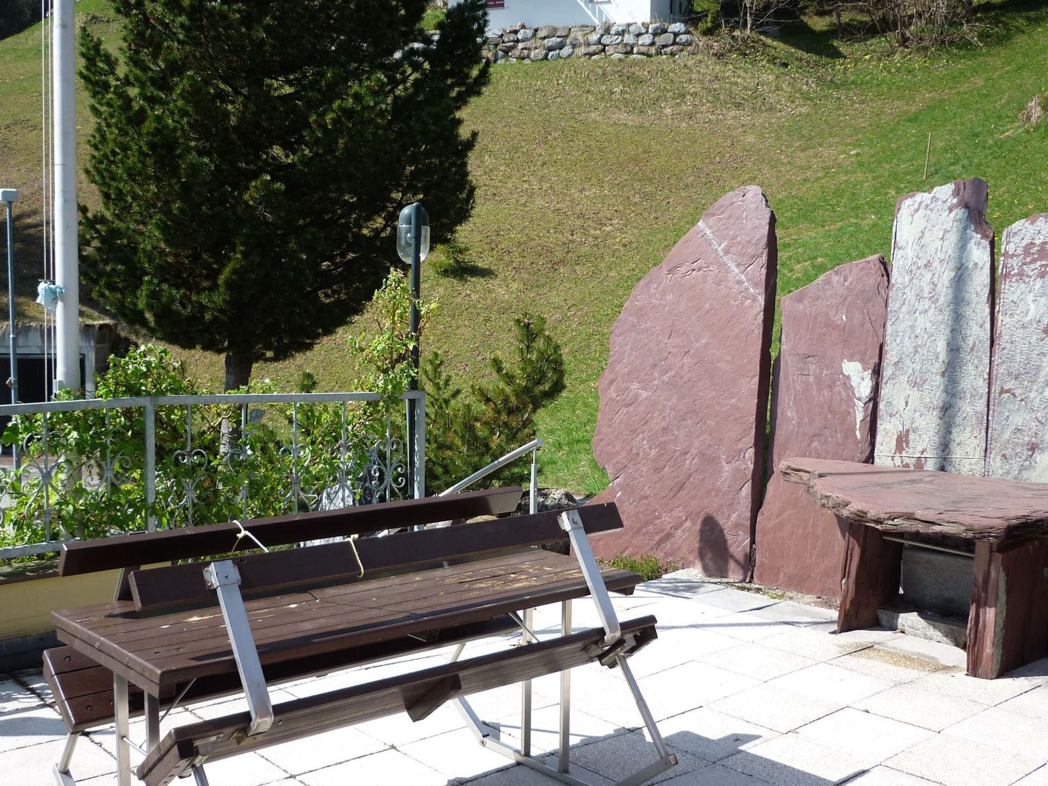 Eine Bank mit Metallgestell, eine Steinmauer und ein grasbewachsener Hügel im Hintergrund.