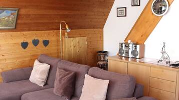 Ein gemütlicher Loft-Raum mit Holzbalkendecke, einem violetten Sofa mit Kissen, einem Holzschrank, einer Uhr und gerahmten Bildern an der Wand.