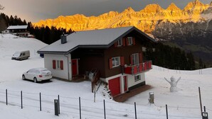 Eine zweistöckige Chalet mit roter und weißer Fassade, einem Balkon und Blick auf verschneite Berge im Abendlicht.