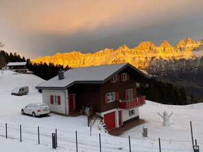 A two-story chalet with a red and white facade, a balcony, and a view of snow-covered mountains at sunset. - Ferienhaus Tschudiboden, (Flumserberg Tannenheim), by Interhome (Flumserberg Tannenheim)