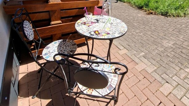Eine Terrasse mit einer Holzbank, zwei Tischen mit Mosaikplatten und einem Grill, mit Blick auf eine gebirgige Landschaft.