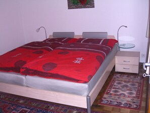 A bed with a red blanket, a bedside table with a lamp, a small drawer, and a patterned rug on the floor. - Ferienhaus Tschudiboden, (Flumserberg Tannenheim), by Interhome (Flumserberg Tannenheim)