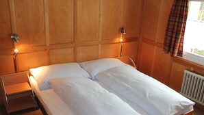 Ein Doppelbett mit weißer Bettwäsche, ein Holz-Kopfteil, Nachttischlampen und ein Fenster mit Vorhängen.