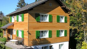 Ein zweistöckiges Holzhaus mit grünen Fensterläden, einer Terrasse mit Gartenmöbeln und einer umliegenden Naturlandschaft.