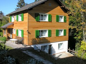 Ein zweistöckiges Holzhaus mit grünen Fensterläden, einer Terrasse mit Gartenmöbeln und einer umliegenden Naturlandschaft.