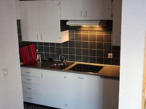 Een kleine keuken met witte kasten, een zwart betegelde spatwand, een roestvrijstalen spoelbak en een rode afdruiprek.