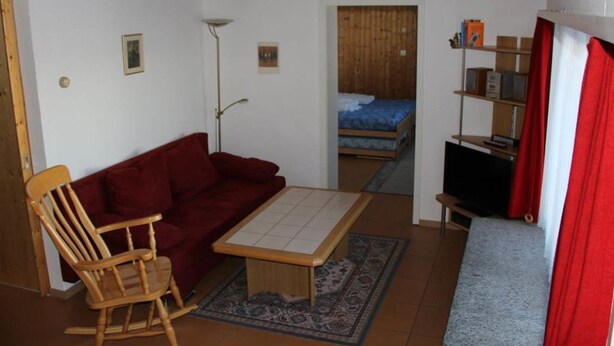 Un espace de vie compact comprenant un fauteuil à bascule en bois, un canapé rouge, une petite table, un téléviseur et un lit à l’arrière-plan.