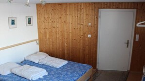 Un lit simple avec une couvre-lit bleue à motifs et des oreillers blancs, une tête de lit en bois, un mur en bois, une porte blanche et un petit tapis sur le sol.