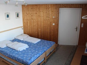 Een eenpersoonsbed met een blauw bedovertrek met patroon en witte kussens, een houten hoofdbord, een houten wand, een witte deur en een klein vloerkleed.