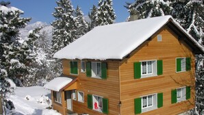 Un chalet de madera de varios pisos con techo cubierto de nieve, rodeado de árboles cargados de nieve y un telón de fondo montañoso.