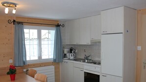 Cocina compacta con armarios blancos, horno empotrado, refrigerador, cafetera y una mesa de comedor con cojines azules.