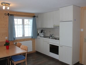 Eine kompakte Küche mit weißen Schränken, einem Einbauofen, einem Kühlschrank, einer Kaffeemaschine und einem Esstisch mit blauen Kissen.