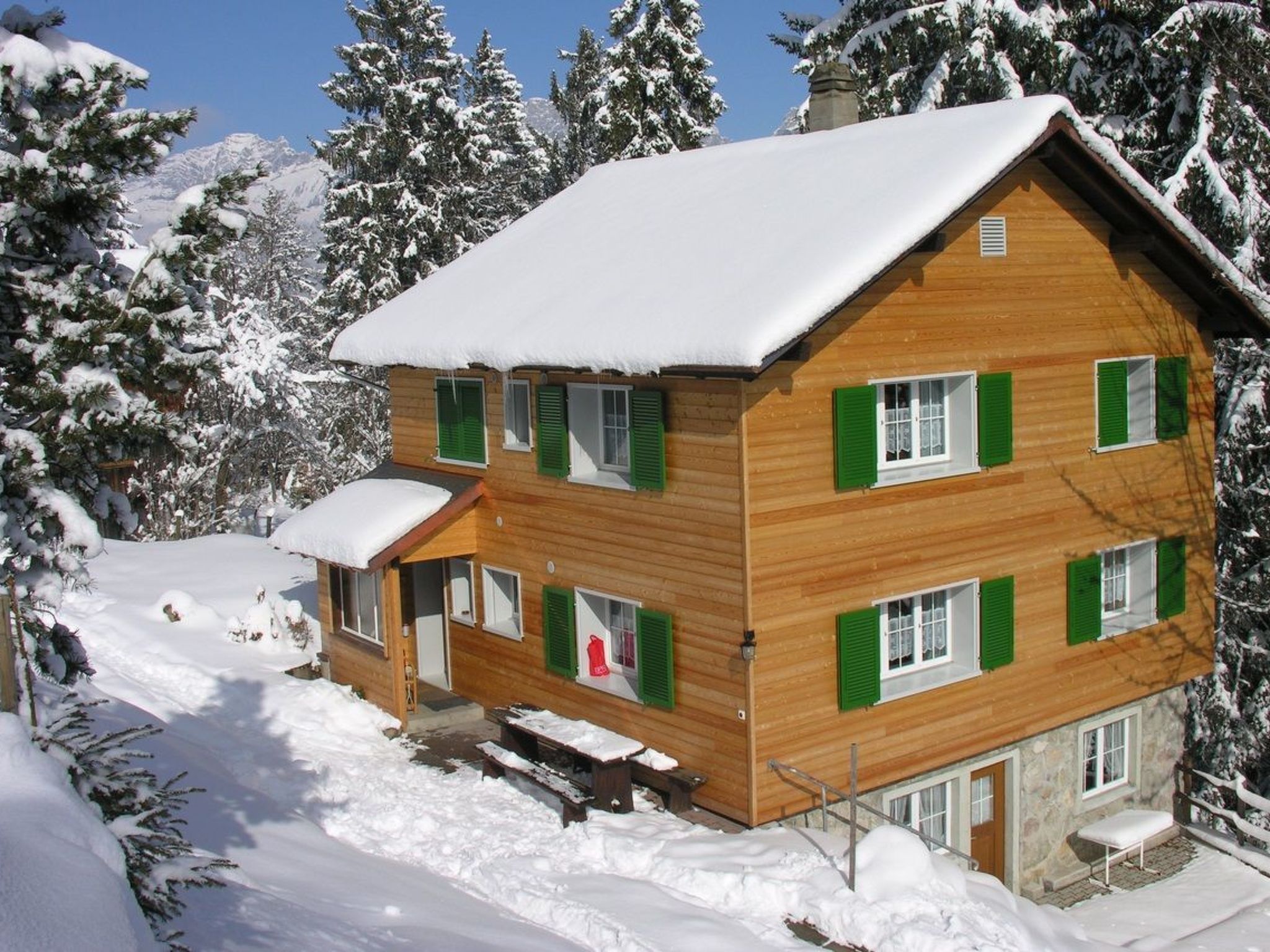 Una cabaña de madera de varios pisos con contraventanas verdes, rodeada de árboles cubiertos de nieve y un cielo despejado.
