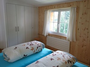 Quarto com duas camas, um guarda-roupa, uma janela com cortinas e painéis de madeira.