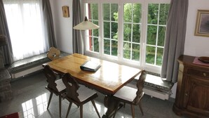 Zona de menjador amb taula i cadires de fusta, finestra amb cortines i una làmpada.