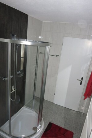 Box de chuveiro envidraçado com piso branco, porta com puxador e toalha vermelha pendurada à direita.