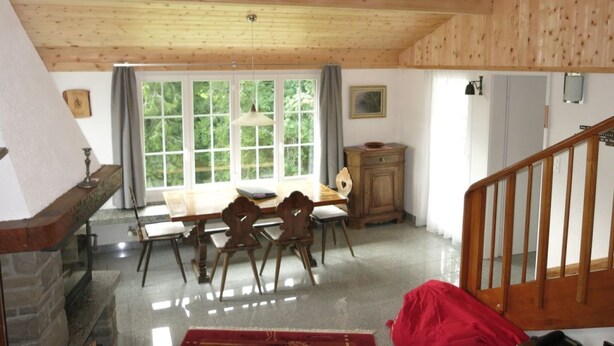 Una habitació amb sostre de fusta, una llar de foc, una taula de dinar amb caires, una finestra amb cortines i una escala.