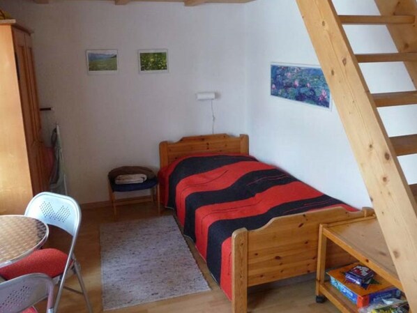 A small, cozy bedroom with a wooden bed, a single chair, a small table, and a ladder.