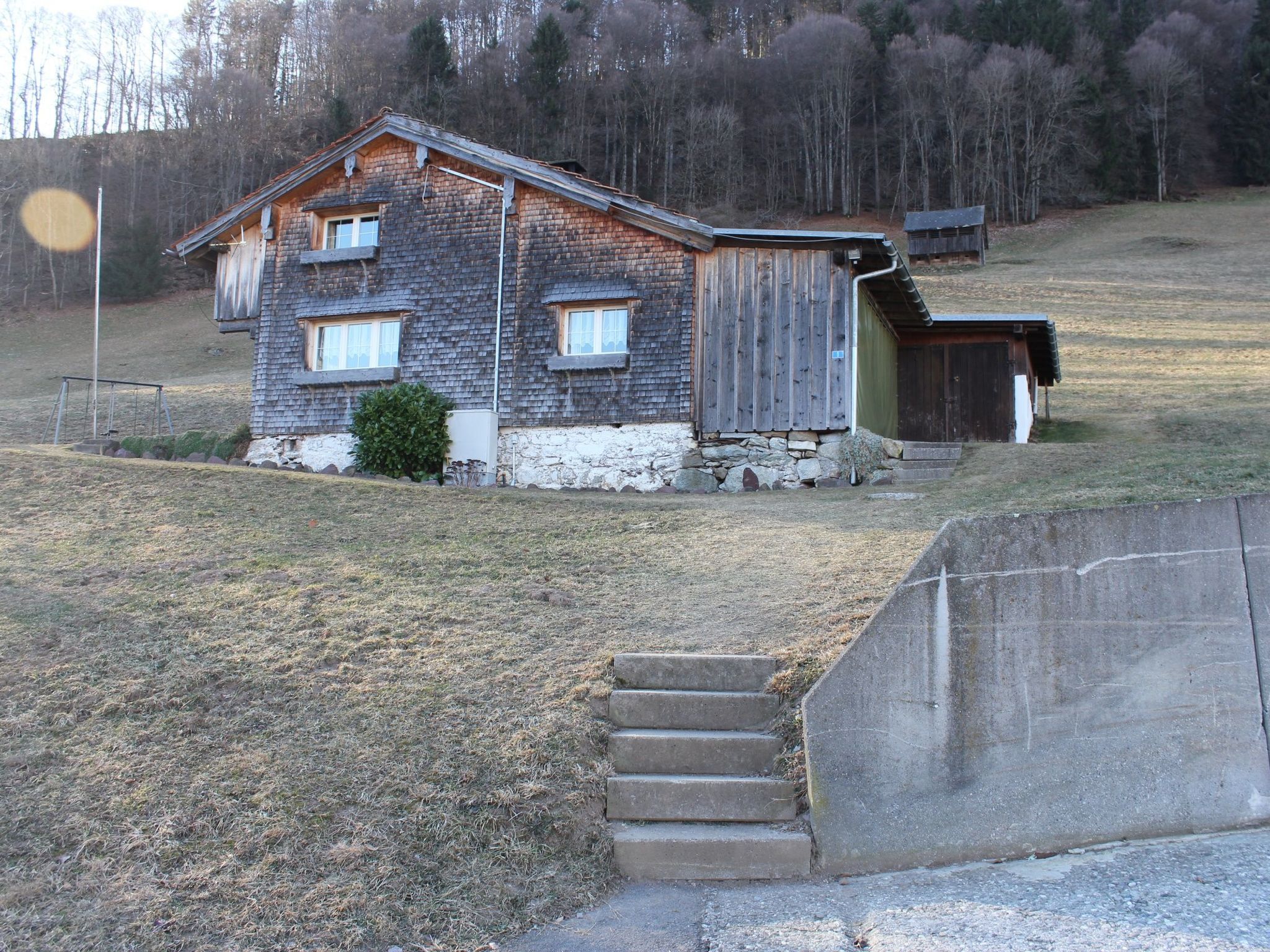 Une maison rustique en bois, avec des fondations en pierre, un petit porche et une rampe en béton menant à l’entrée.