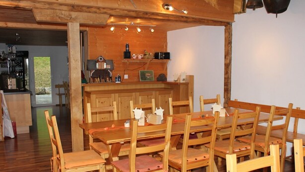 Un coin repas en bois avec une longue table et des chaises, une cuisine en arrière-plan et une vue sur l’extérieur.