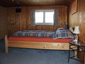Une pièce de cabane en bois comprenant un lit, une fenêtre avec des rideaux, une petite table avec une lampe et un porte-manteau.