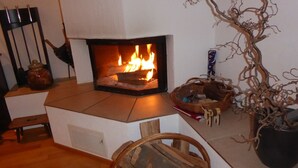 Un camino acceso, una panca in legno, un cesto di legna e una pianta decorativa.