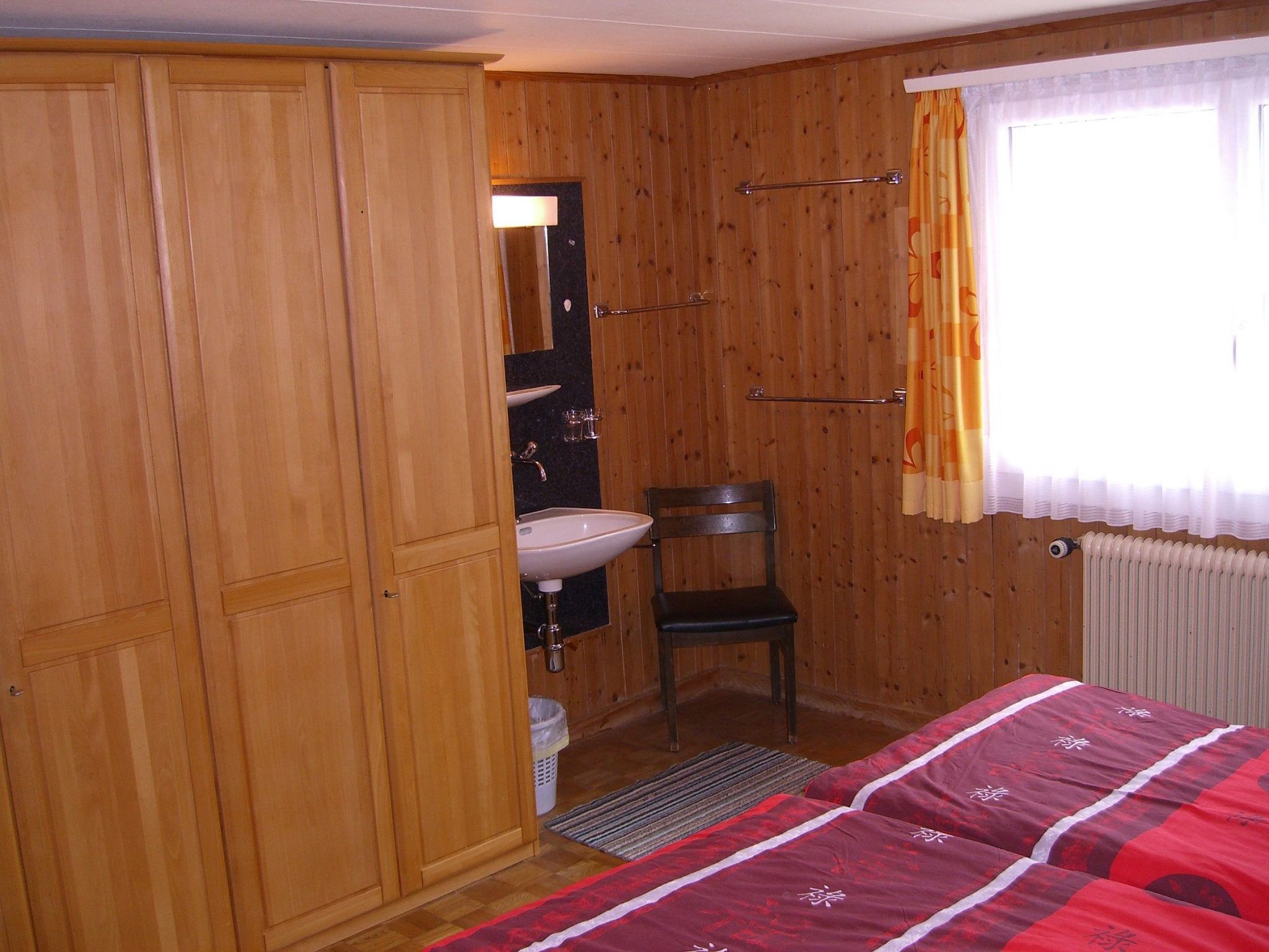 A wooden-paneled room with a bed, a chair, a sink, and a window with curtains.