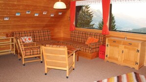 Een gezellige kamer met een houten plafond, een zithoek, een kleintje tafeltje, een raam met gordijnen, en uitzicht op bergen.