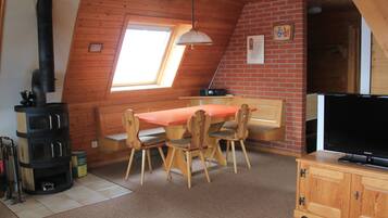 Ein gemütliches Zimmer mit schrägen Wänden, einem Kamin, einem Esstisch mit Stühlen, einem Fernseher und einem kleinen Fenster.