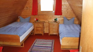 Een kleine, houten slaapkamer onder het dak, met twee bedden, een raam met rode gordijnen en een houten dressoir.