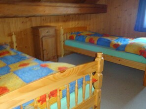 Ein Zimmer aus Holz mit zwei Betten, jedes mit einer farbenfrohen Stegdecke. Es gibt einen Holzschrank und ein Fenster mit einem blauen Vorhang.
